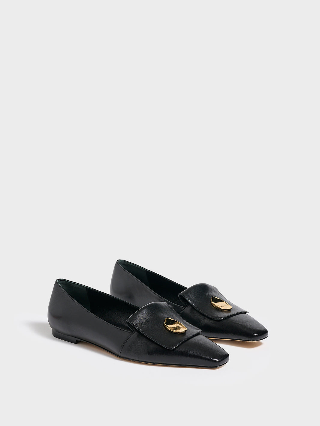 Angle view of a pair of black leather flat  loafers with a  gold hardware detail and squared toe. 